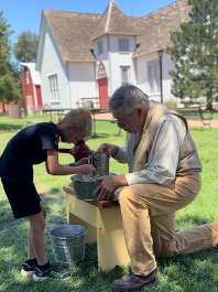 A living history volunteer at the Humphrey Heritage Village assists a youngster to sift flour with a hand-cranked sifter.