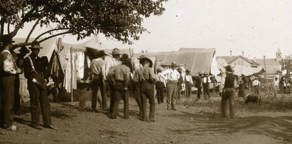 Freedmen gathered and walking along a road at Fort Gibson c. late 1800s. Trees, wagons, and tents are seen well into the distance.