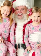 Santa with two children on his lap, all are smiling. The children are wearing Christmas pajamas