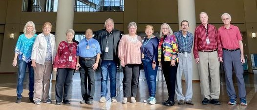 A row of Oklahoma History Center Volunteers and Steve Hawkins stand in the Devon Great Hall underneath a replica of the Winne Mae airplane