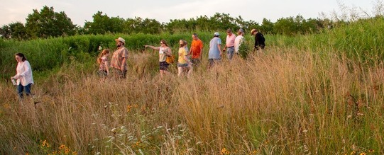 A photo of a group of people talking a walk at the Spiro Mounds Archaeological Site. Wildflowers are growing in the foreground