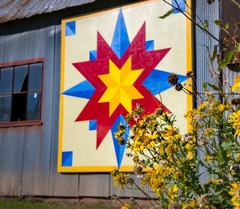 A photo of a barn quilt with vibrant colors, attached to the side of a metal building. Wild flowers are growing in the foreground