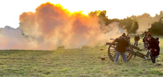 Reenactors fire cannons at sunset, a cloud of smoke above the cannons is taking the color of the sky