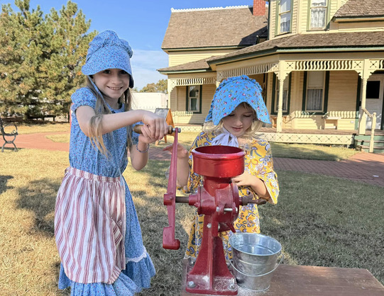 Two schoolchildren dressed in pioneer clothing, work to grind corn with a hand-cranking machine on a table near historic home