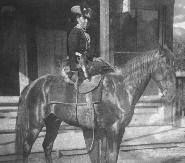Belle Starr seated side saddle on horseback with a holster and pistol on her left hip, she has hold of the reigns and a riding whip