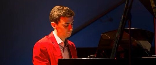 Nathan Avakian seated at the piano in a red jacket