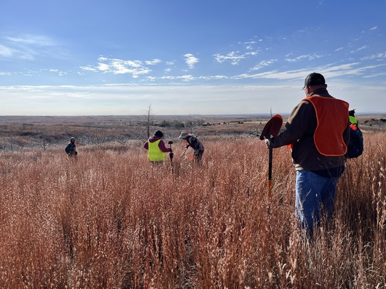Surveying the Battle of Turkey Springs site. Several archaeologists, surveyors are standing in a field of tall grass with fluorescent vests