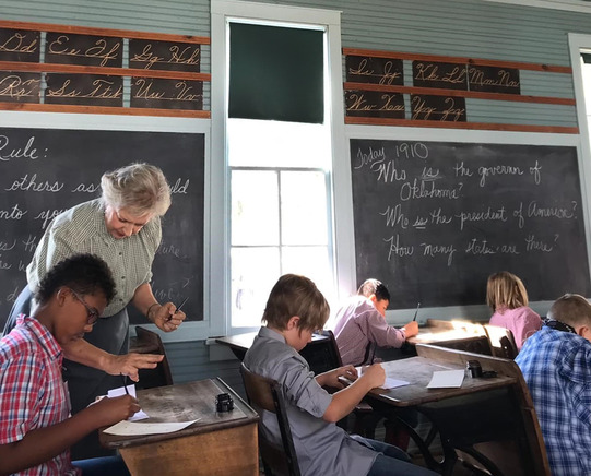A volunteer schoolmarm teaches students how to write in pen and ink. Students are seated at wooden desks writing with quills