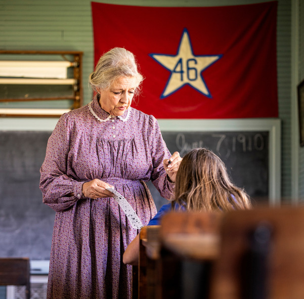 A schoolmarm stands in front of the chalkboard at Turkey Creek School instructing a student