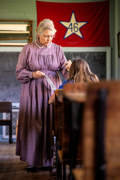 A schoolmarm stands in front of the chalkboard at Turkey Creek School instructing a student