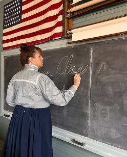 A schoolmarm writes in chalk on the chalkboard of the Turkey Creek Schoolhouse. An American flag hangs above