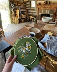 An Embroidery hoop and the lap of the person working on the project with a fireplace hearth and logs of the cabin in the background