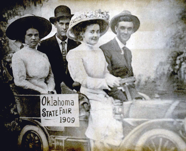 Two couples, in period dress from 1909, seated in a prop of a cardboard car, posing for a group photo at the Oklahoma State Fair