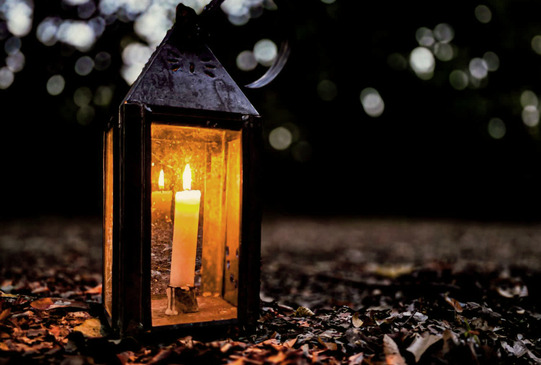 A candle lit inside a glass lantern sits on the trail in Doaksville with leaves around it