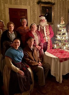 Living history volunteers and staff dressed for Christmas, pose around a small fir tree decorated for the season.