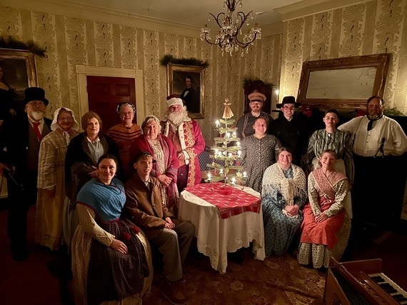 Living history volunteers and staff dressed for Christmas, pose around a small fir tree decorated for the season.