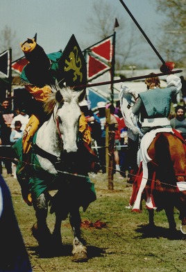 Two men dressed in medieval era clothing joust on horseback.
