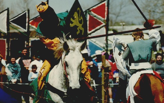 Two men dressed in medieval era clothing joust on horseback.