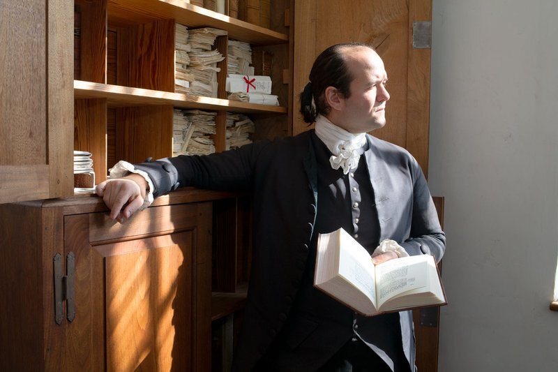 A living history actor playing James Madison rests his right arm on shelving, with a book in hand.