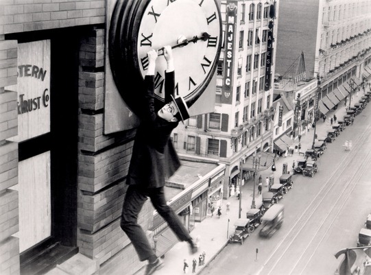 Screen capture from the film Safety Last. The scene features actor Harold Lloyd dangling from large hands of a clock over downtown Los Angeles.
