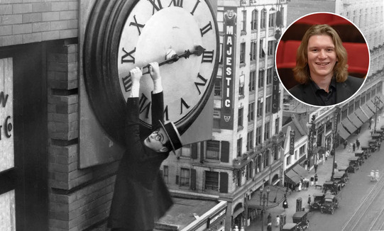Harold Lloyd hangs from the hands of a large clock several stories up while traffic and people pass below.