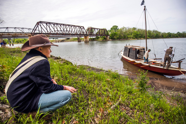 A young man wearing historical clothing, kneels at the Three Forks river system, observing men operating a keelboat.