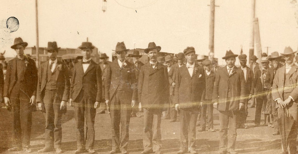 Seven men standing in a row in double breasted coats, vests, suits, ties, and hats outside the Perry Oklahoma train depot.