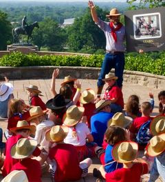 Bart Taylor stands in front of a group of children seated in a half circle around him. They are all wearing cowboy gear.