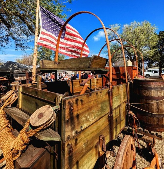 An American flag flies behind a wooden buckboard wagon with staves, as a crowd gathers