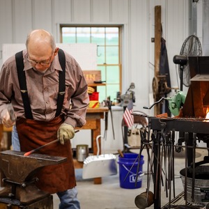 Tom Nelson hammers a piece of red hot iron at the anvil, with a roaring fire in his blacksmith forge to his left.