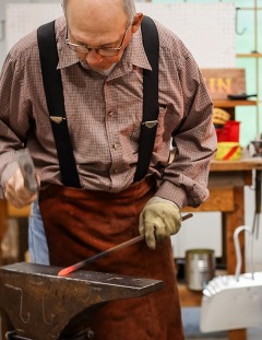 Tom Nelson hammers a piece of red hot iron at the anvil, with a roaring fire in his blacksmith forge to his left.