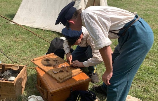 Two living history reenactors work together at a demonstration of troop preparation. They are wearing wool caps and pants