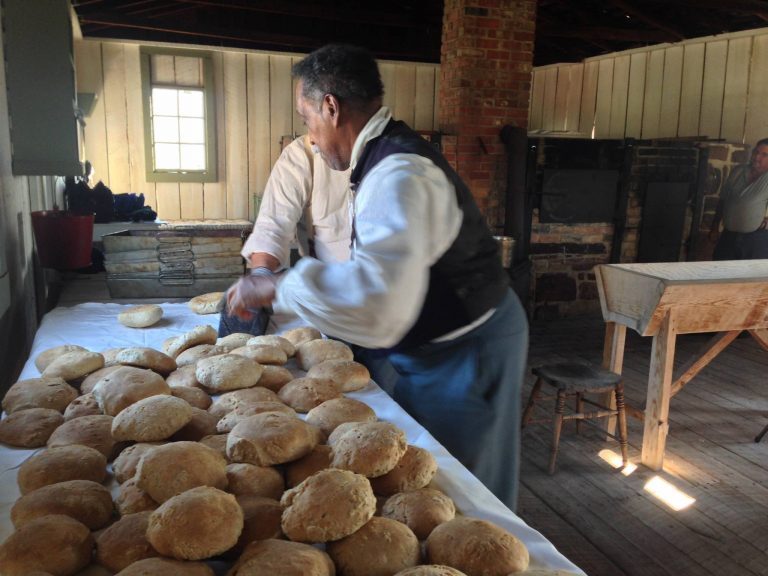 Men dressed in Union uniforms and aprons sort newly baked loaves of bread at the Fort Gibson bakehouse onto tables.