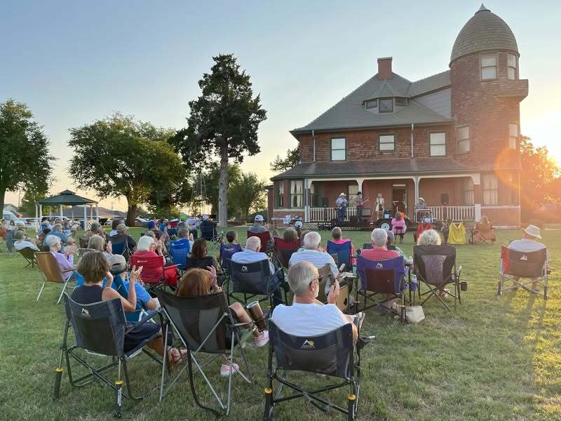 A group of people sitting in lawn chairs listen to a concert on the steps of an historic home as the sun is setting.