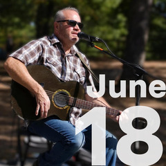 Travis Kidd wears sunglasses, and is seated playing the guitar for a crowd outdoors.