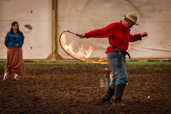 A roper performs a trick at a Wild West Show with a fiery lasso as an onlooking perform looks on.