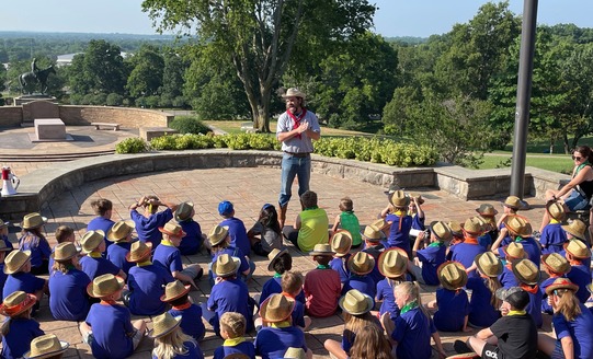 Young children gather at the steps of the Will Rogers Memorial Museum wearing cowboy hats, listening to a group leader dressed in Western Gear.