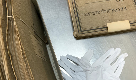 White gloves, an antique book, and other archived files are placed on a museum archives work table.