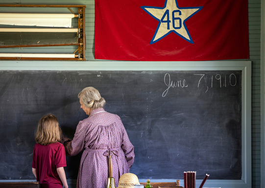 A schoolmarm teaches writing and penmanship to a young student as they stand at a chalkboard of a one-room school.