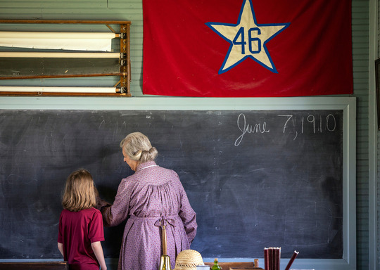 A schoolmarm teaches writing and penmanship to a young student as they stand at a chalkboard of a one-room school.