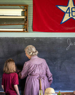 A schoolmarm teaches writing and penmanship to a young student as they stand at a chalkboard of a one-room school.