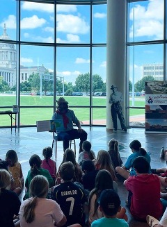 A man dressed as a cowboy, seated in front of a crowd of schoolchildren, reads a book for story time at the Oklahoma History Center.