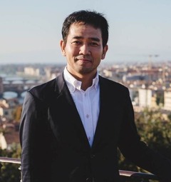 A man dressed in a black suit stands on a balcony with an Italian city skyline, possibly Florence.