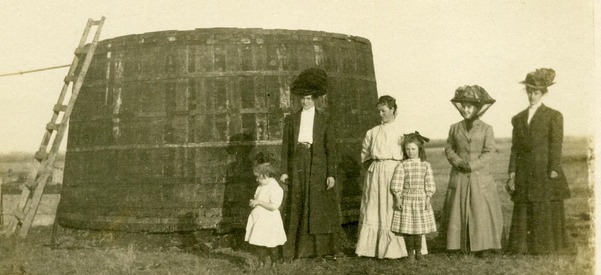 Four women and two girls standing next to a large oil storage container shaped like a large barrel, with a 10 ft. ladder propped on its side.