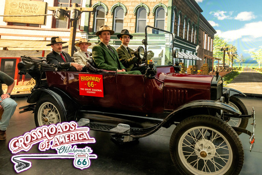 A roadster with men and women seated inside, situated in front of a green screen displaying a large building.