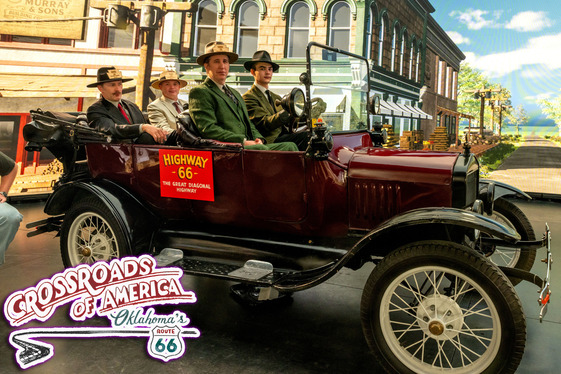 A roadster with men and women seated inside, situated in front of a green screen displaying a large building.