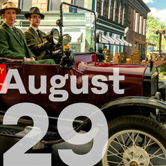 Two men driving an antique car look to the camera with buildings on a green  screen in the background. Large lettering August 29