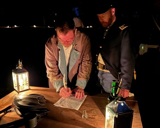 Two men dressed in historical clothing look at and sign a document laying on a table flooded with candlelight.