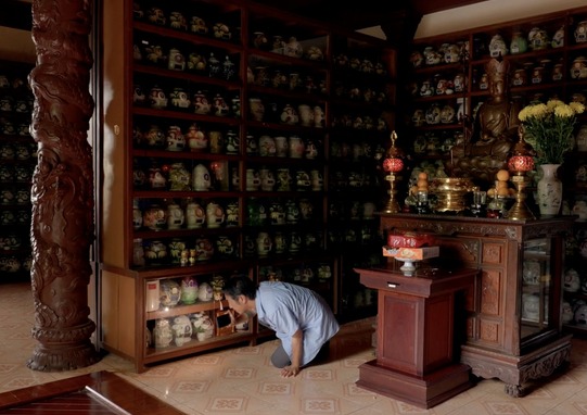 A man on his hands and knees holds his cell phone flashlight to a shelf of ceramic vessels on multilayered shelves.
