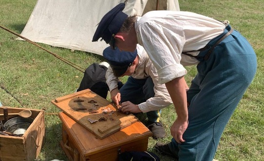 Two soldiers dressed as Infantrymen, hover over maps and equipment near an encampment.